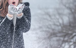Woman in cozy winter clothing blowing snowflakes with excitement outdoors in a snowy setting.
